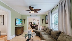A living room with a sofa, a coffee table, and a ceiling fan. There is a dining area in the background with a table and chairs. - Bahama Breeze Bungalow (West Palm Beach)