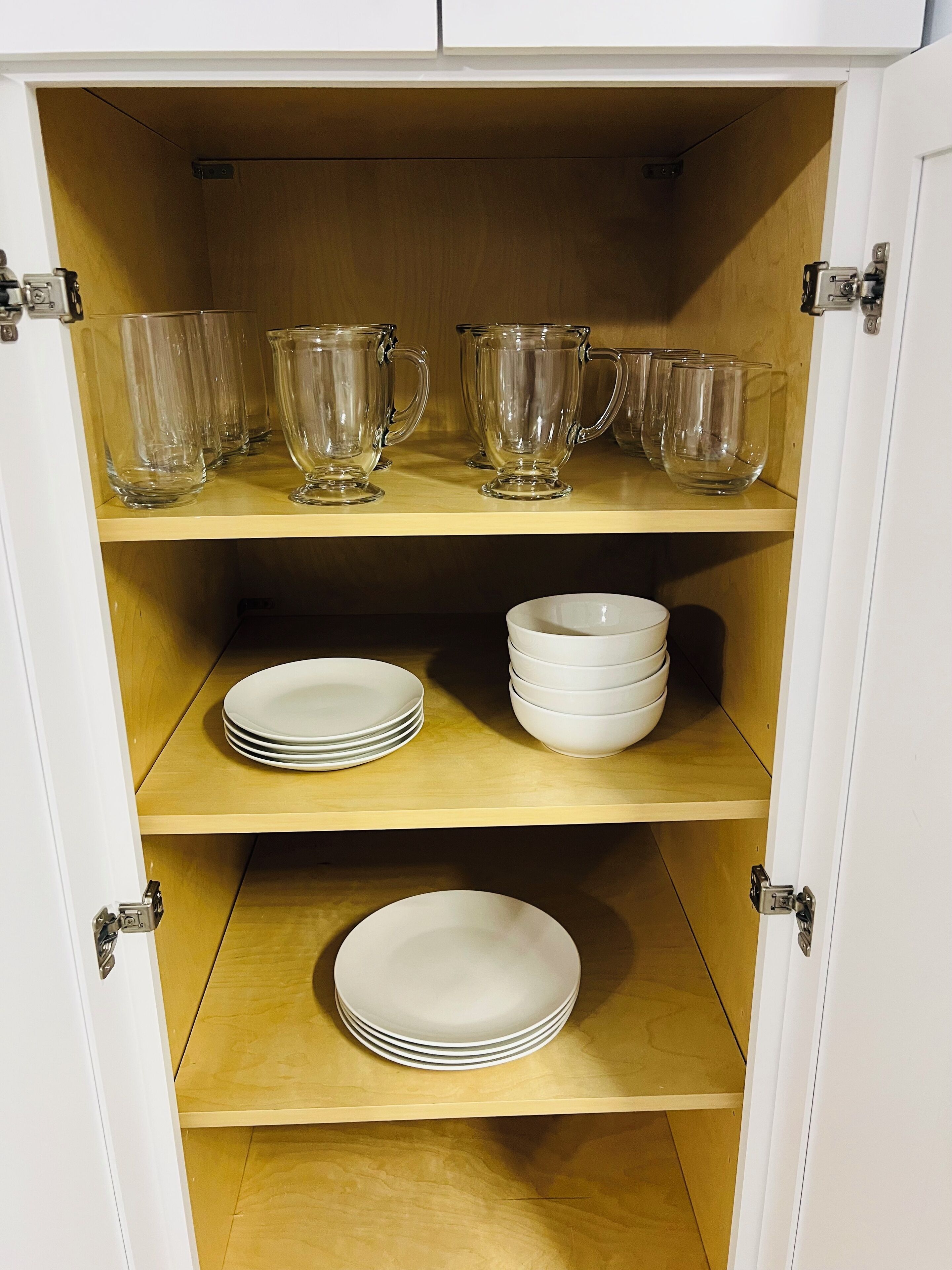 A cabinet with three shelves; the top shelf holds clear glass pitchers, the middle shelf has white plates, and the bottom shelf also has white plates.