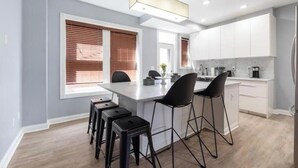 A modern kitchen with white cabinets, a central island with bar stools, and a window with blinds. - Luxury Chefs Kitchen - OSU/Short North - Sleeps 10 (Columbus)
