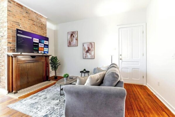 A living room with a brick fireplace, a gray sofa, and a flat-screen TV mounted on the wall.