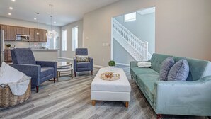 Ein großzügiges Wohnzimmer mit einem grünen Sofa, zwei Sesseln, einem Couchtisch und einem Korbwäschekorb. Im Hintergrund ist eine Treppe zu einem Balkon zu sehen.