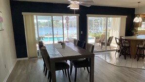 Ein Essbereich mit großem Tisch und Stühlen, ein Küchenbereich mit einer Bar und große Schiebeglasfenster, die zu einem Pool führen.