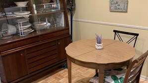 A dining area with a round wooden table and chairs, a wooden cabinet with glass display shelves, and a wall-mounted shelf with decorative items.