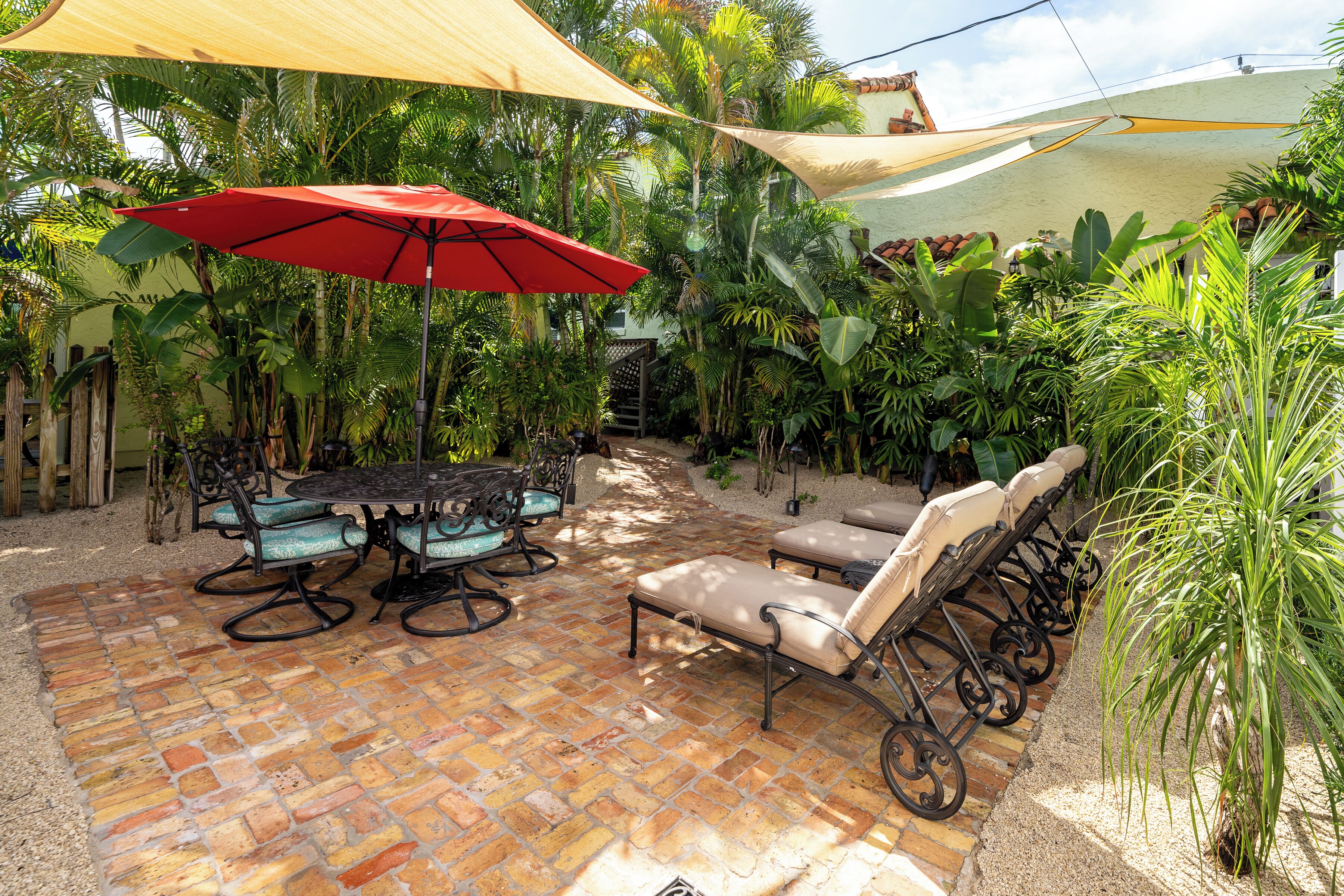 A patio with a brick floor, a table with chairs, a red umbrella, and lounge chairs.