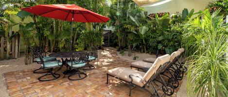 A patio with a brick floor, a table with chairs, a red umbrella, and lounge chairs.
