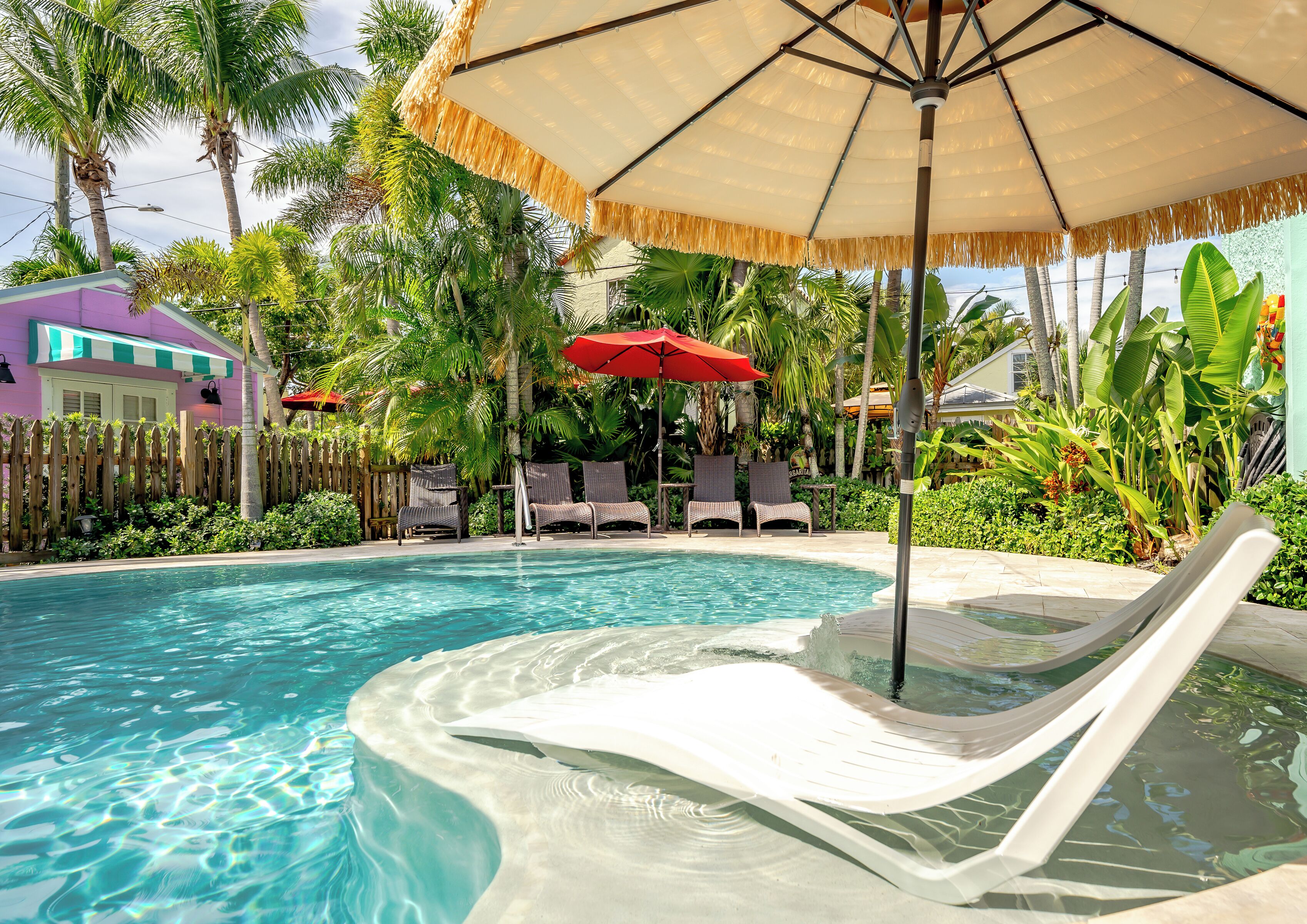 A swimming pool with lounge chairs and umbrellas, surrounded by tropical plants and a wooden fence.
