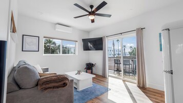 A modern living room with a ceiling fan, a flat-screen TV, a sofa, a coffee table, and a balcony with outdoor seating.