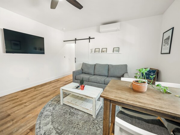 A modern living room with a grey sofa, a white coffee table, a wooden dining table, and a ceiling fan.
