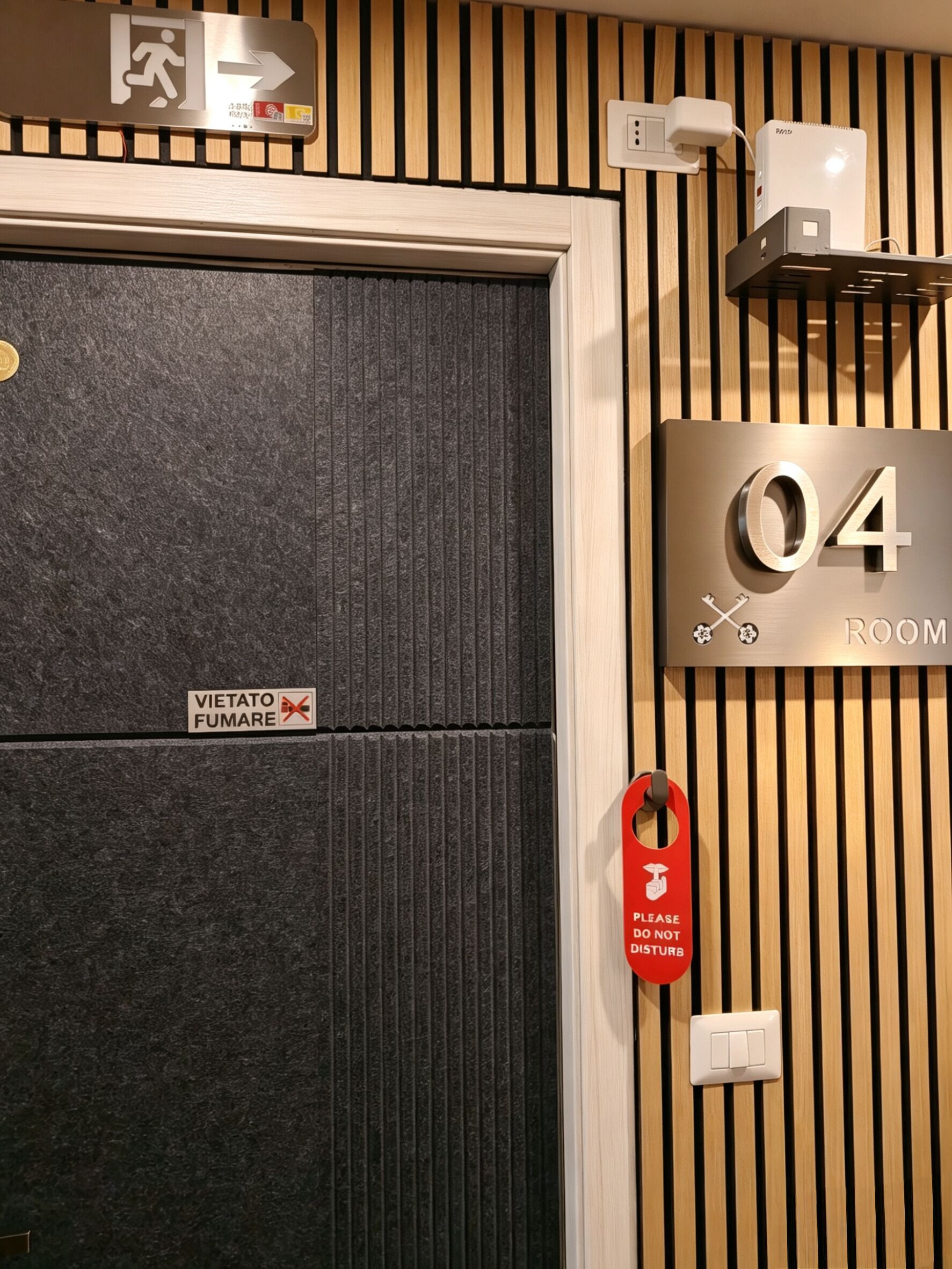 A hotel room with a door labeled 'Vietato Fumare,' a number '04' sign, a red 'Please Do Not Disturb' sign, and a power outlet.