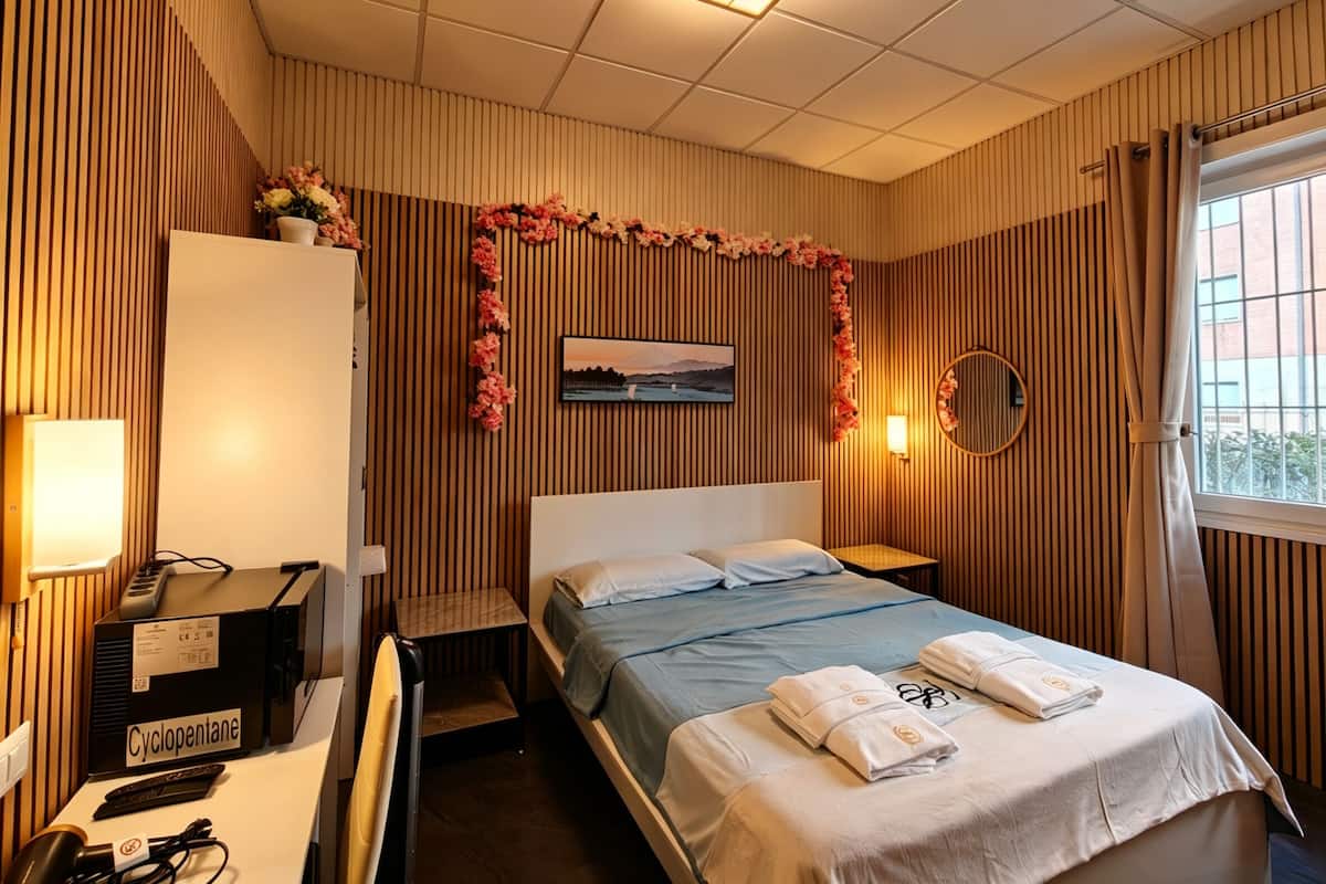 A hotel room with a bed, a desk with a computer, a lamp, and a window with curtains.