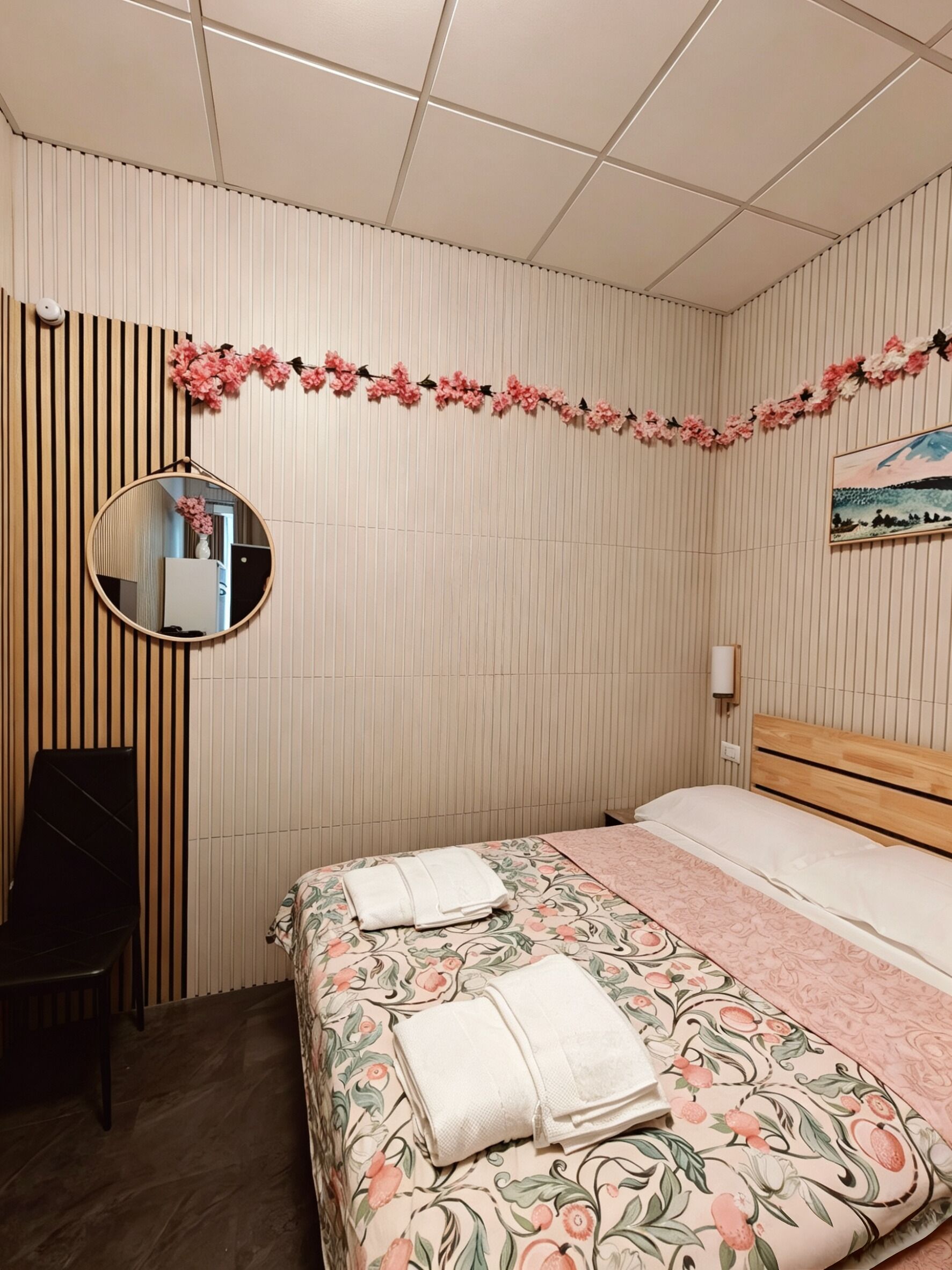 A bedroom with a bed, a round mirror, a wooden chair, and a wall-mounted light fixture.
