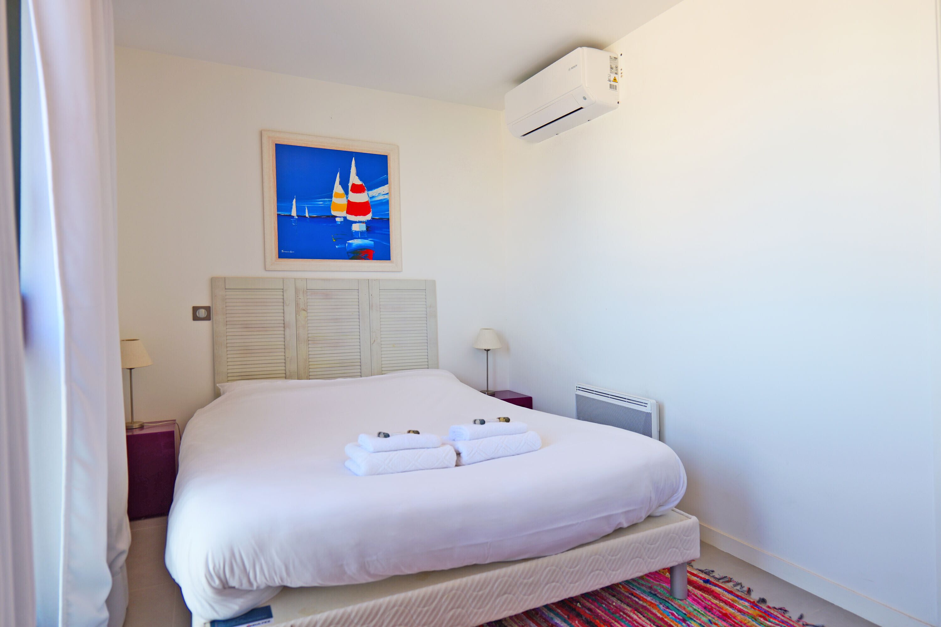Una cama bien hecha con sábanas blancas y dos toallas, un cuadro de veleros en la pared y un equipo de aire acondicionado sobre la cama.