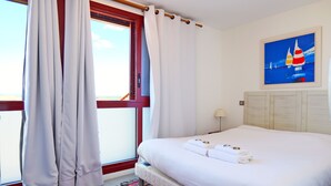 Una habitación de hotel con una cama grande, cortinas blancas, un tapete colorido y un cuadro de veleros en la pared.