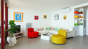Una sala moderna con un sofá blanco, sillas rojas y amarillas, una mesa de centro y un televisor. Hay cuadros coloridos en la pared y una planta en maceta.