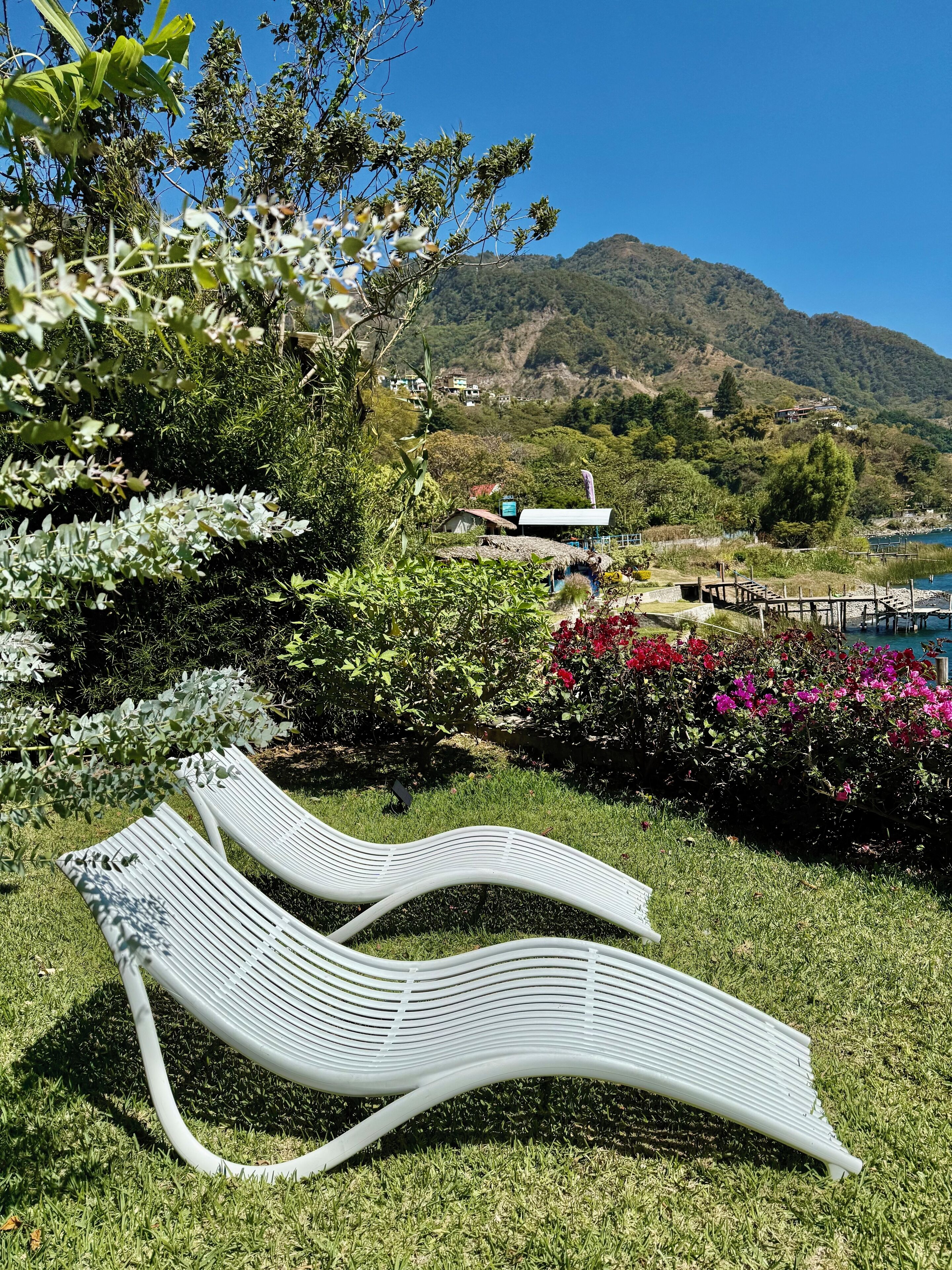 Two white lounge chairs in a garden with a view of a mountain and a building in the background.