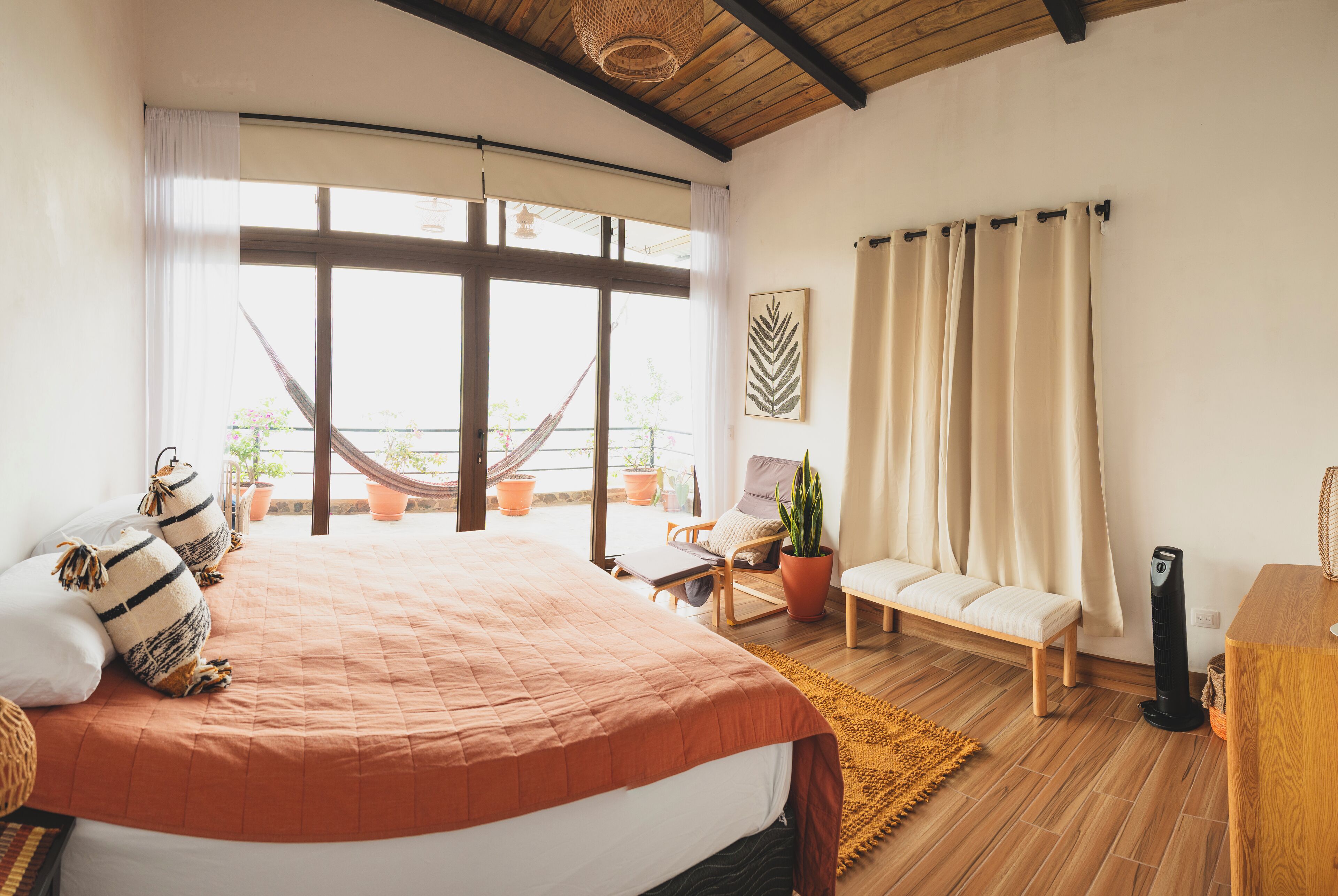 Une chambre avec un grand lit, un hamac, une chaise, une plante en pot et une vue sur l’extérieur.