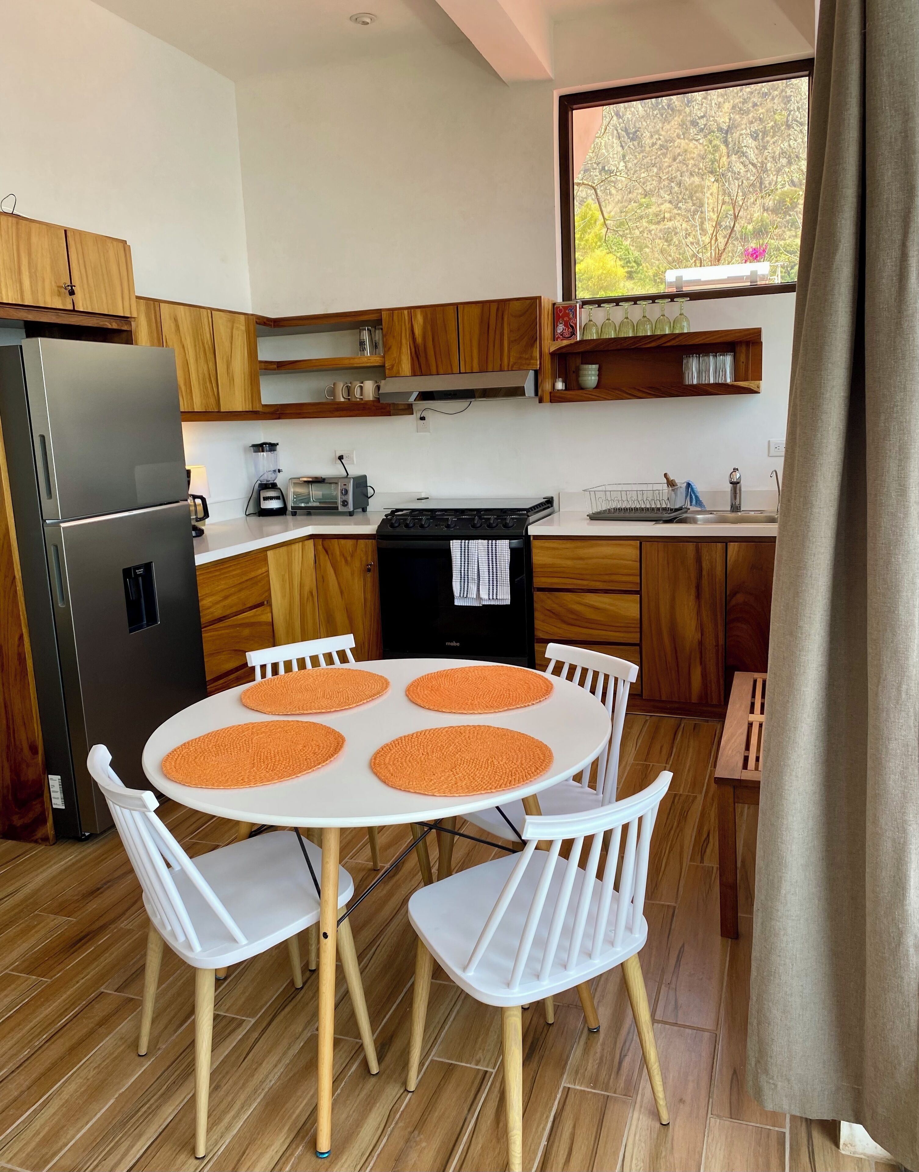 Une cuisine moderne avec des meubles en bois, un réfrigérateur en acier inoxydable, une cuisinière et un coin repas comprenant une table blanche et des sets de table orange.
