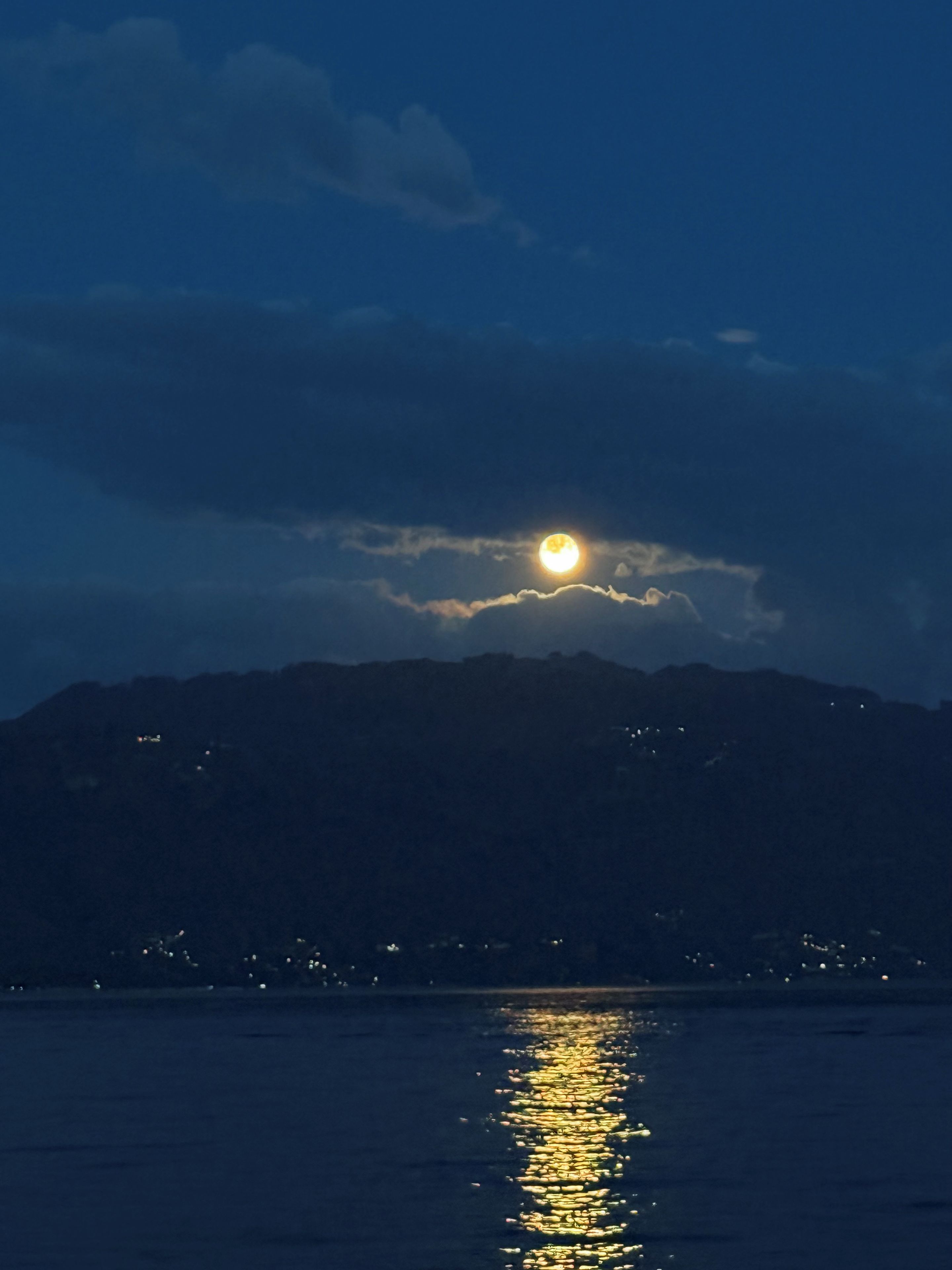 Une scène nocturne avec la lune se reflétant sur l’eau, des montagnes en arrière-plan et des lumières lointaines.