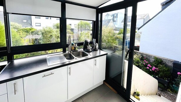 A modern kitchen with white cabinets, a stainless steel sink, and a view of a garden and buildings through large windows.