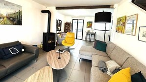 A modern living room with a sofa, armchair, and a small wooden table. There is a fireplace with a chimney, a dining area with a table and chairs, and artwork on the walls.