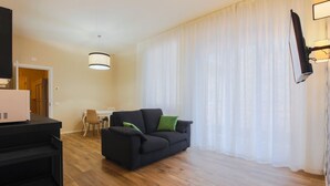 Un salon avec un canapé noir, une table à manger avec des chaises, un téléviseur à écran plat fixé au mur et un luminaire suspendu au plafond.