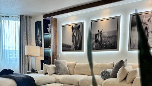 Un salon moderne avec un canapé d’angle, une table basse et un tapis. Des tableaux encadrés ornent le mur, et une lampe est posée sur une table d’appoint.