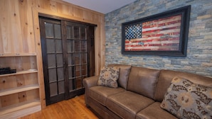 Een kamer met een houten deur, een bruine leren bank met gestreepte kussens, en een Amerikaanse vlag in een lijst op een stenen muur.