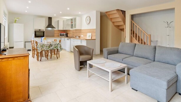 A modern living room with a grey sofa, a coffee table, and a dining area with a table and chairs. There is a kitchen in the background with cabinets and appliances.