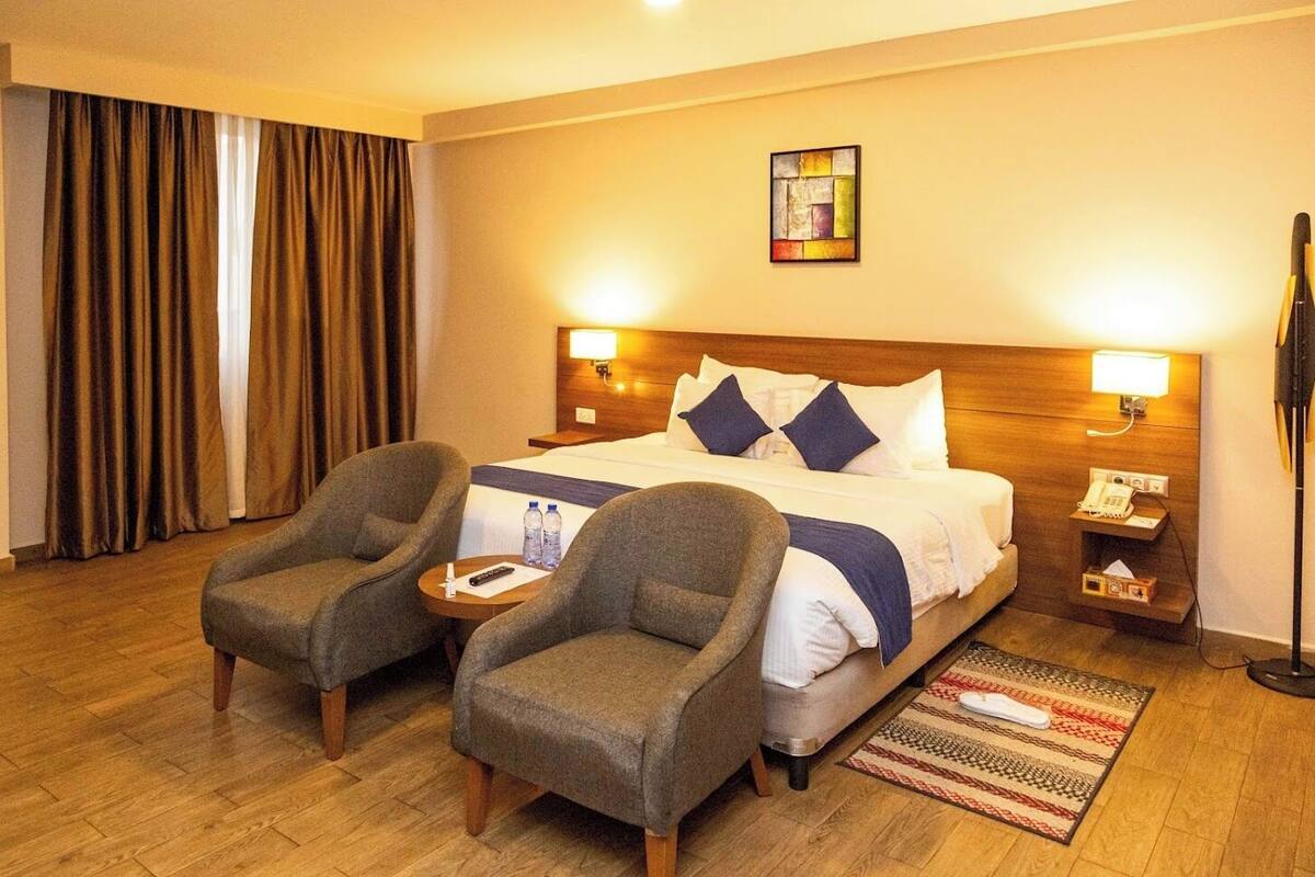 A hotel room with a bed, two armchairs, a small table with a remote control, a phone, and a lamp.