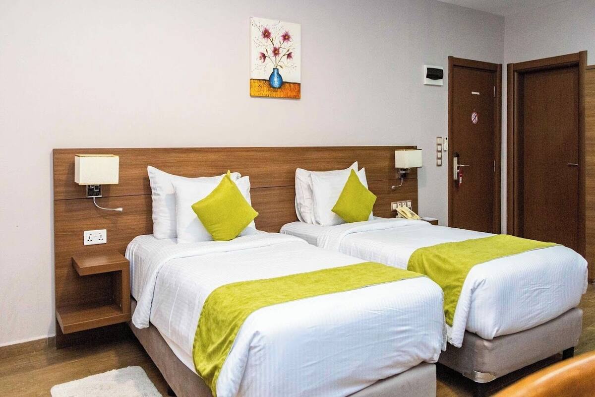 A hotel room with two beds, each with white bedding and green throw pillows, a wooden headboard, bedside lamps, a small bedside table, and a wall-mounted artwork.