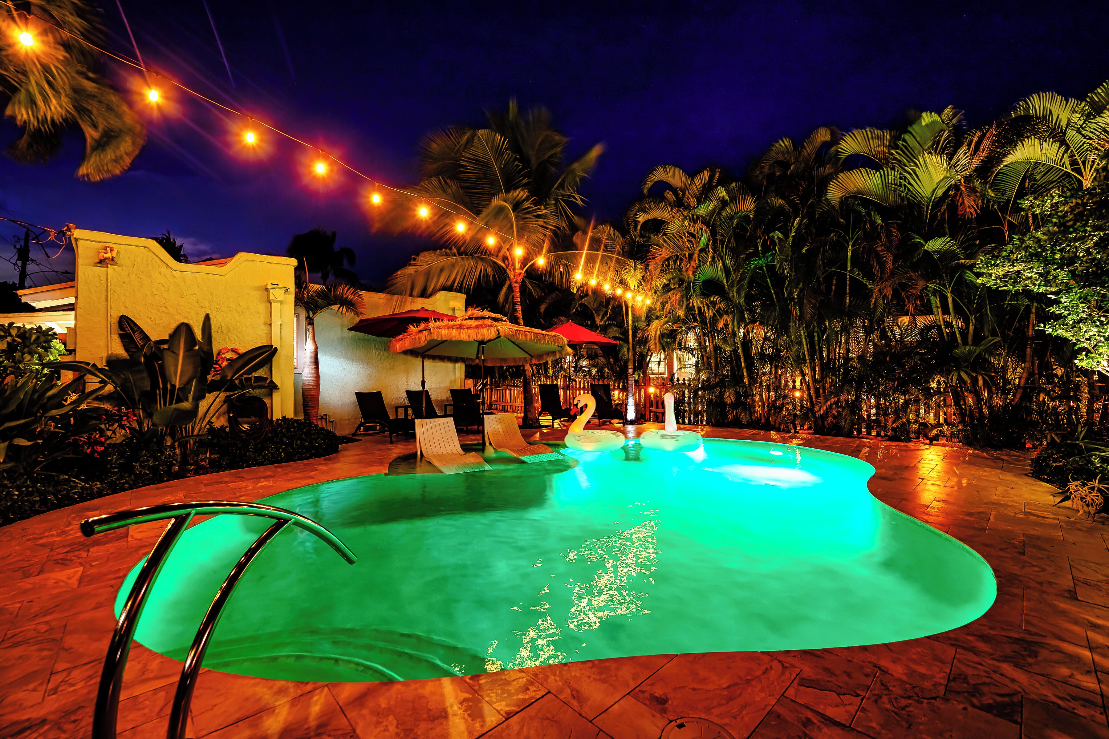 A swimming pool with a green-lit edge, surrounded by palm trees and outdoor seating.