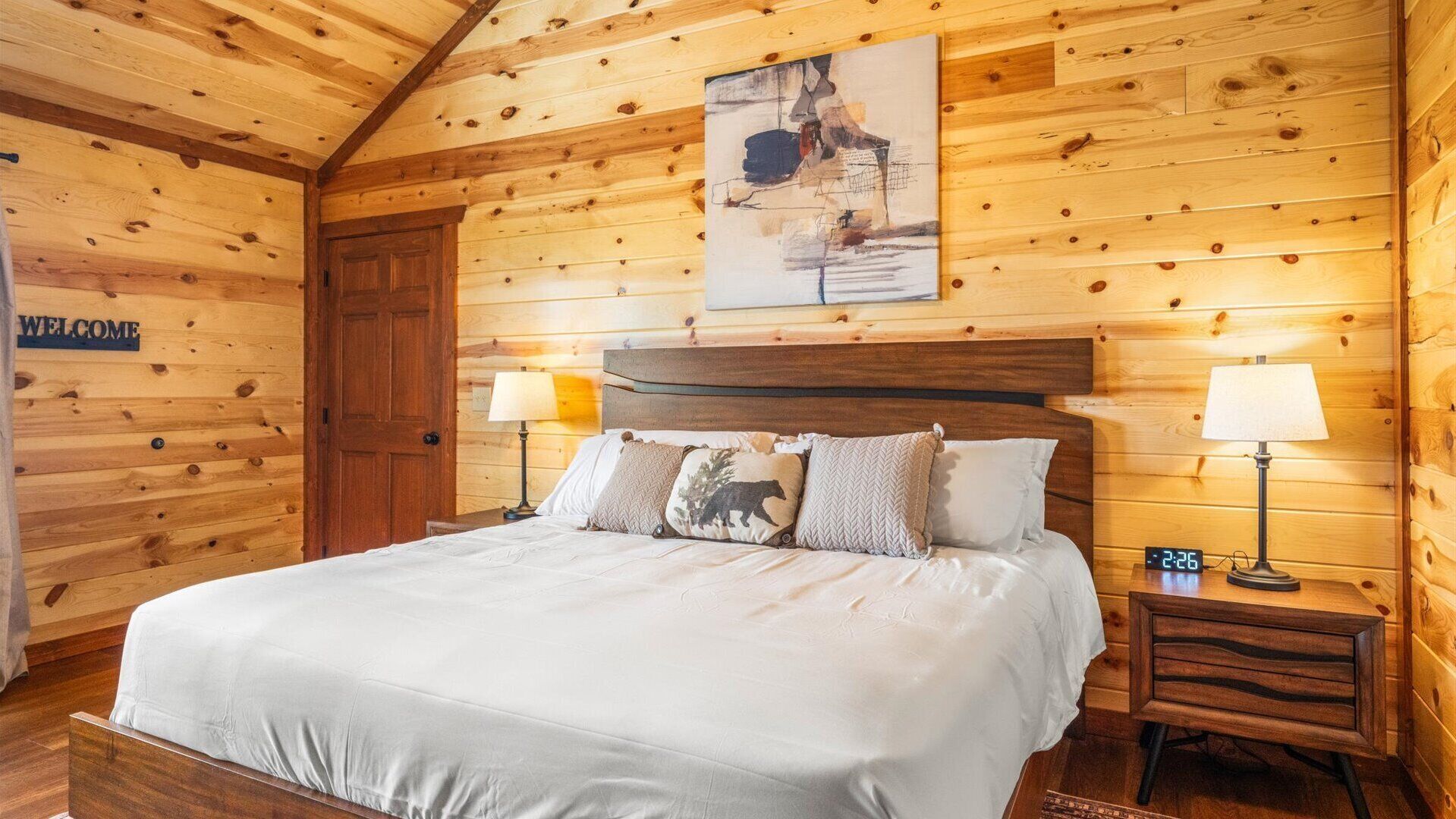 Una habitación de cabaña de madera con una cama, dos lámparas de noche, un reloj y un letrero de bienvenida.