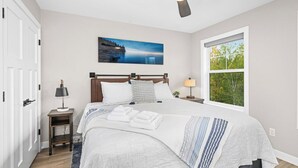 A bedroom with a bed, a nightstand, a ceiling fan, a window, and a painting on the wall.