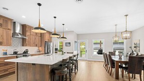 A modern kitchen with wooden cabinets, a large island with bar stools, pendant lighting, and a dining area with a large table and chairs.