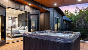 A modern outdoor hot tub area with a wooden deck, a glass-enclosed bedroom, and a view of a cabin.