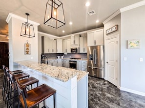 A modern kitchen with white cabinets, a granite island, bar stools, stainless steel appliances, and pendant lighting.