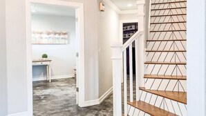 Un pasillo con piso de concreto, una escalera con escalones de madera y un diseño geométrico en la pared, y una puerta que da a una habitación con un escritorio y una planta.