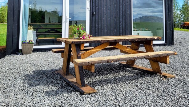 A modern, dark wooden cabin with large glass doors and windows, a wooden picnic table, and a gravel ground.