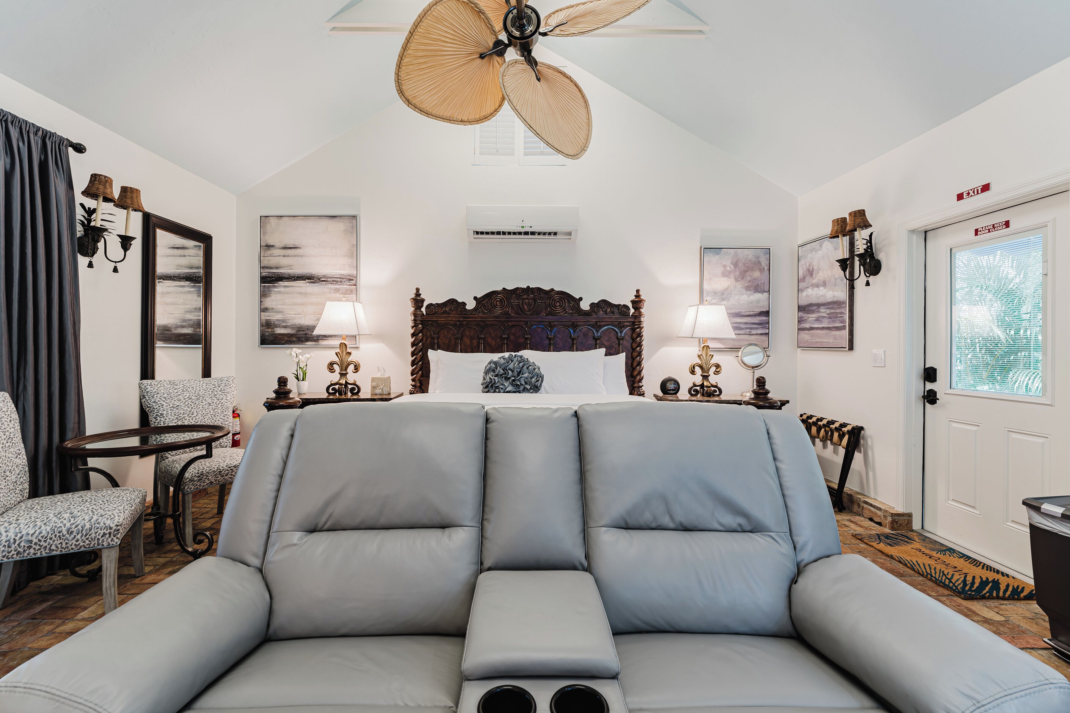 A spacious room with a large grey sofa, a ceiling fan, a bed with a wooden headboard, and artwork on the walls.