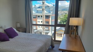 A hotel room with a large bed, a wooden nightstand, a table with a lamp, and a view of buildings and mountains through the window.