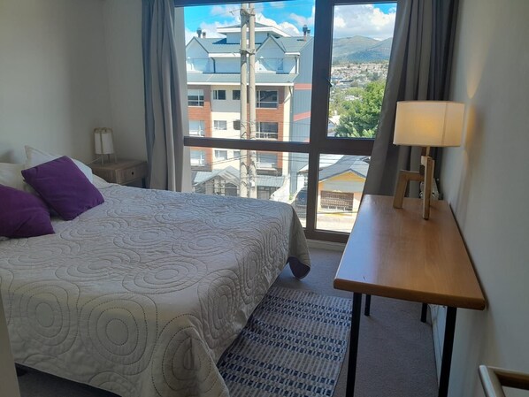 Habitación de hotel con una cama grande, una mesita de noche de madera, una mesa con lámpara y vistas a edificios y montañas por la ventana.