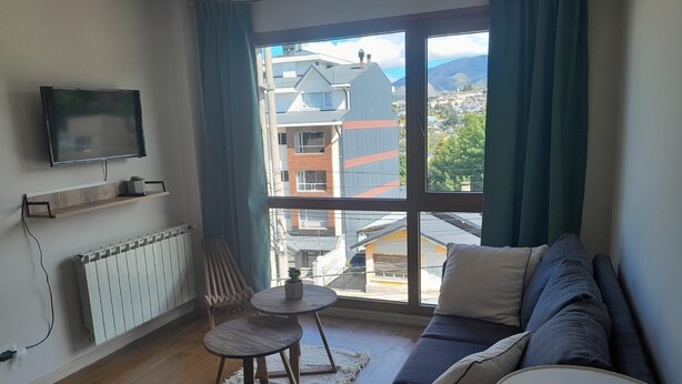 A modern living room with a large window, a sofa, a small round table, and a TV mounted on the wall.