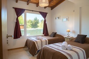 Ein Schlafzimmer mit zwei Betten, einem Fenster mit Vorhängen, einem Nachttisch mit Lampe und Blick auf Bäume.