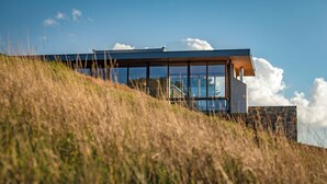 Un edifici modern amb grans finestres de vidre, situat sobre un turó verd amb cel serè.