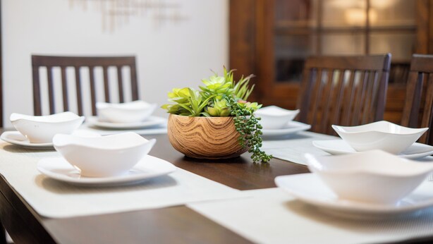 A dining table set with white bowls and plates, a wooden bowl with green plants, and wooden chairs.