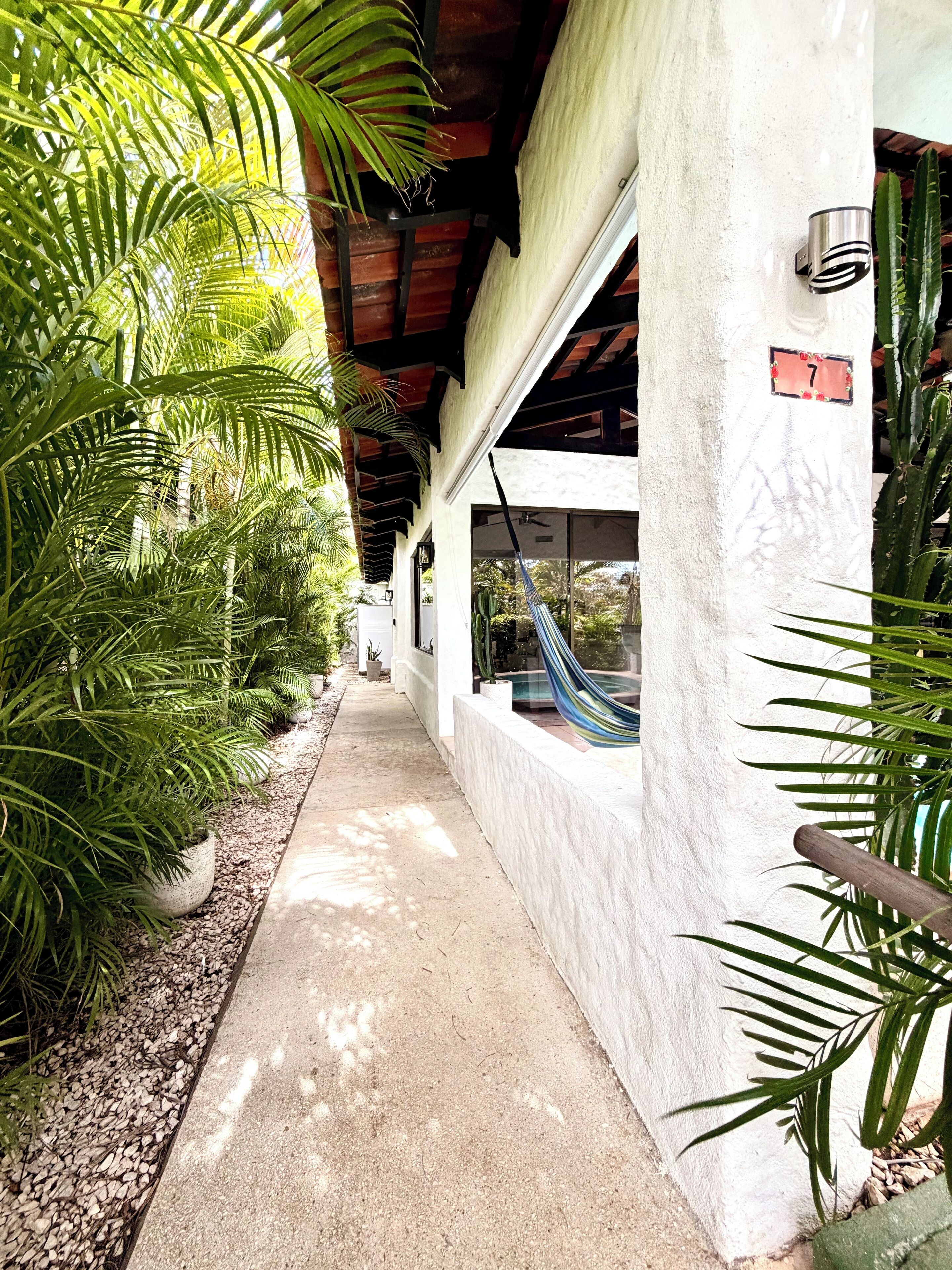 A pathway lined with potted plants leading to a building with a white exterior and a number sign '7'.