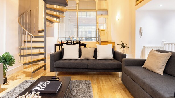Een moderne woonkamer met een grijze bank, houten vloeren, een trap en een groot raam met jaloezieën.