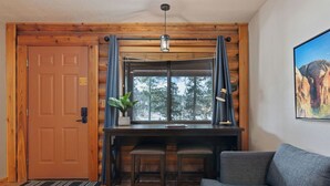 A cozy room with a wooden desk, two chairs, a grey sofa, and a large window with curtains.