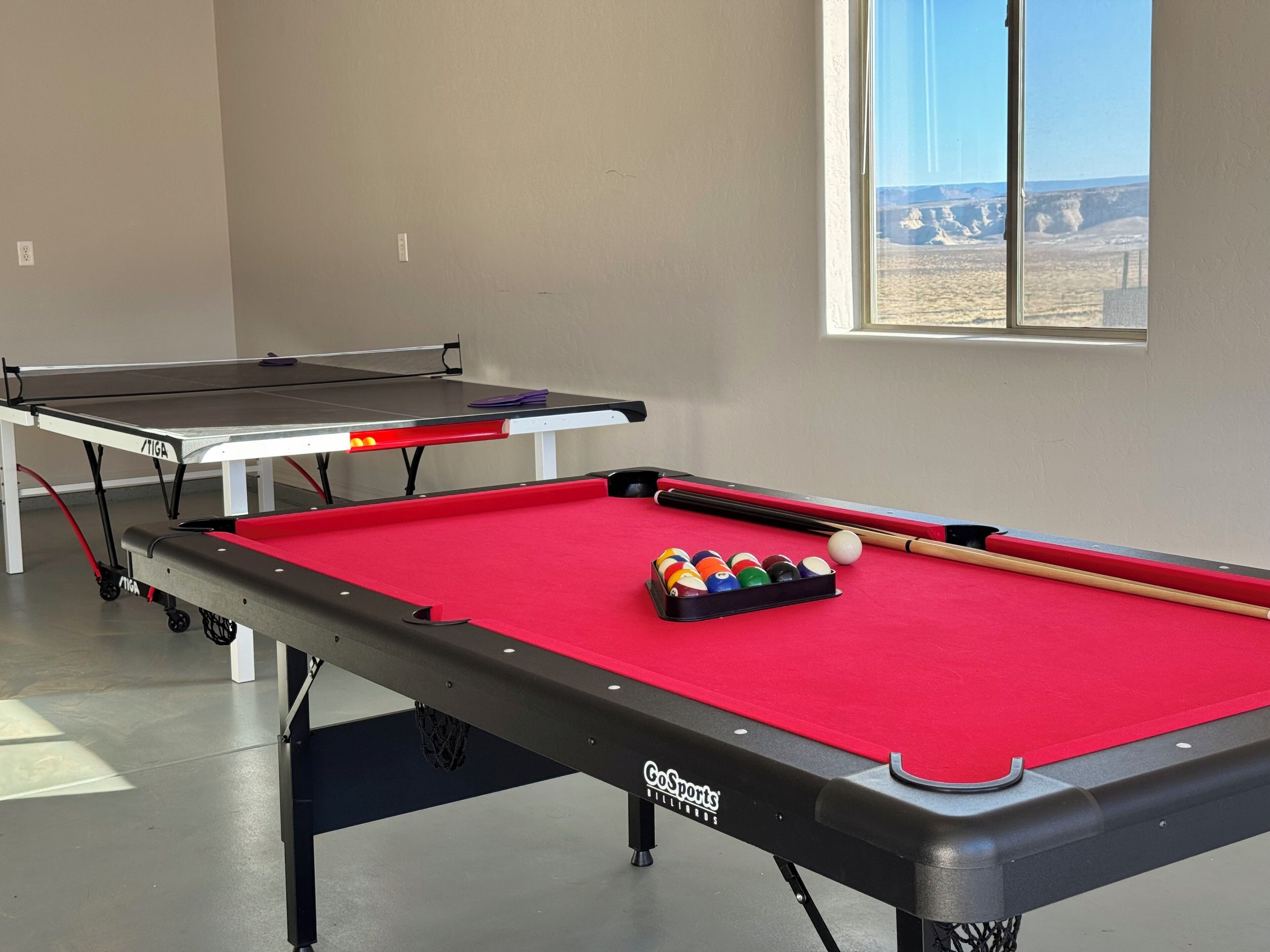 Une table de billard avec des billes et des queues, une table de ping-pong, et une fenêtre donnant sur un paysage désertique.