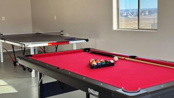Une table de billard avec des billes et des queues, une table de ping-pong, et une fenêtre donnant sur un paysage désertique.